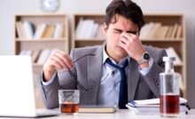 A young man in a business suit drinks alone at his desk, to indicate Recognizing the Signs of a High Functioning Alcoholic