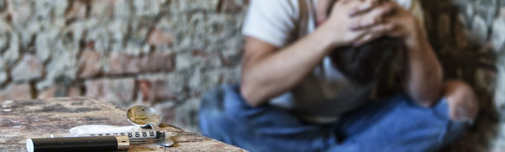 Heroin Detox - A needle and some white powder sit on a table. In the background slightly out of focus a man sits with his legs crossed and his hands on his head. He is addicted to heroin and needs a heroin detox to get clean so he can start rehab.