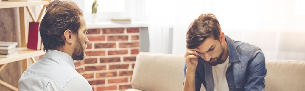 The Continuum of Care - An addiction treatment counselor sits with his back to the camera as he discusses the best level of treatment as outlined by the Continuum of Care. His male patient sits on the couch with his head down thinking about his responsibilities and goals for his treatment.